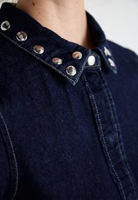 Camicia in denim blu navy con una consistenza morbida, colletto strutturato e bottoni a pressione metallici lungo il colletto e la placca frontale.