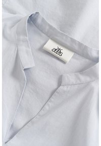 Camicia in lino azzurro chiaro con scollatura a V e un'etichetta che mostra un logo. Il tessuto appare morbido e leggermente texture, con un colletto rilassato.