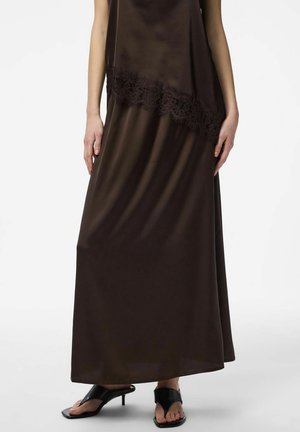 Žena v dlhých tmavohnedých saténových šatách s čipkovým zdobením a čiernymi otvorenými sandálmi na opätku, stojaca na bielom pozadí.