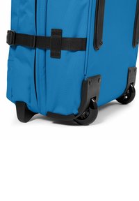 Valigia in tessuto blu con base in plastica nera e assemblaggi delle ruote; presenta una superficie piatta e una cinghia in tessuto per fissare il contenuto.