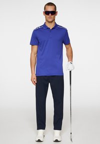 Royal blue polo shirt with white stripes on shoulders, navy pants, white golf shoes, and large purple sunglasses, holding a golf club.