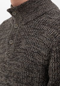 Grå och brun texturerad stickad tröja med hög krage och tre knappar. Nära vy som visar ribbat tyg och knappdetaljer.