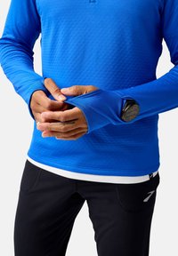 Blaues langärmliges Sportshirt aus strukturiertem Stoff, mit Daumenlöchern an den Ärmeln und einem weißen Unterlayer, der am unteren Ende sichtbar ist. Passende schwarze Sporthose.