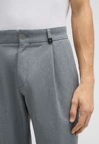 Pantaloni grigi su misura realizzati in tessuto testurizzato, con chiusura a bottone e una leggera piega sul davanti. Un'etichetta nera è visibile sulla vita.