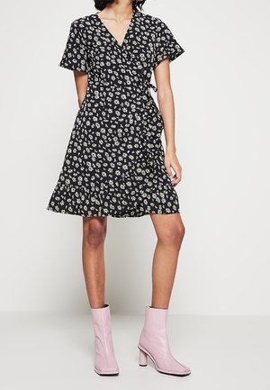 Femme portant une robe noire portefeuille imprimée de fleurs blanches et des bottines roses pâles, debout devant un fond clair uni.