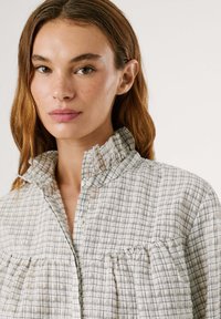 Karirana, lahka srajca z nabranim ovratnikom in gumbnim zapiranjem. Ima mehak material v nevtralnih tonih in nabran detajl na prsih.