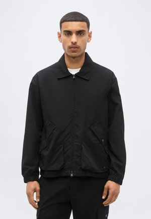 Mann mit kurzen Haaren, der eine schwarze Jacke mit Reißverschluss, Fronttaschen und elastischen Bündchen trägt, steht vor einem einfarbigen hellgrauen Hintergrund.