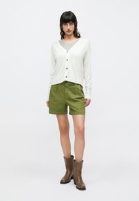 Λευκό μακρυμάνικο ζακέτα με μαύρα κουμπιά συνδυασμένο με πράσινα σορτς. Το μοντέλο φοράει καφέ μποτάκια μέχρι τον αστράγαλο· λείες υφές και άνετη εφαρμογή.