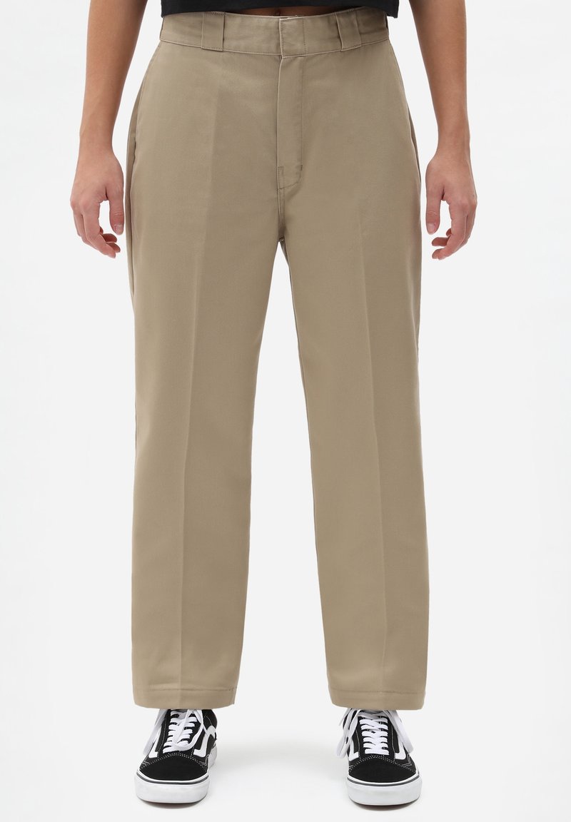 Personne portant un pantalon beige à jambes droites et des baskets noir et blanc, debout devant un fond blanc uni.