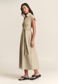 Bež haljina na kopčanje s remenom u struku, kratkim rukavima i lepršavim dizajnom do koljena. Nošena s crnim sandalama. Glatka tekstura tkanine.