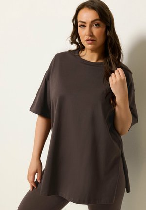 Kvinde med langt mørkt hår iført løs mørkegrå kortærmet skjorte og matchende bukser, poserer mod en ensfarvet lys væg.