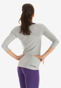 Grå langærmet top med lilla detaljer, tætsiddende design og en kant med logoet "Winshape." Kombinationen med lilla leggings.