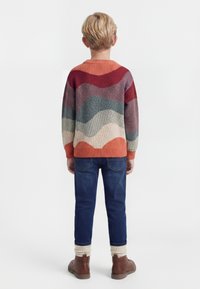 Enfant aux cheveux blonds vu de dos, portant un pull à motifs ondulés multicolores, un jean bleu, des chaussettes crème et des bottes marron.