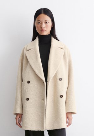 Cream-colored double-breasted coat in textured fabric with large lapels and dark buttons, featuring side pockets and relaxed fit.