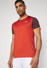 Nike urheilupaita, jossa on punainen vartalo, tummat hihat ja oranssit yksityiskohdat. Valmistettu hengittävästä kankaasta, siinä on pyöreä kaula-aukko ja lyhyet hihat.