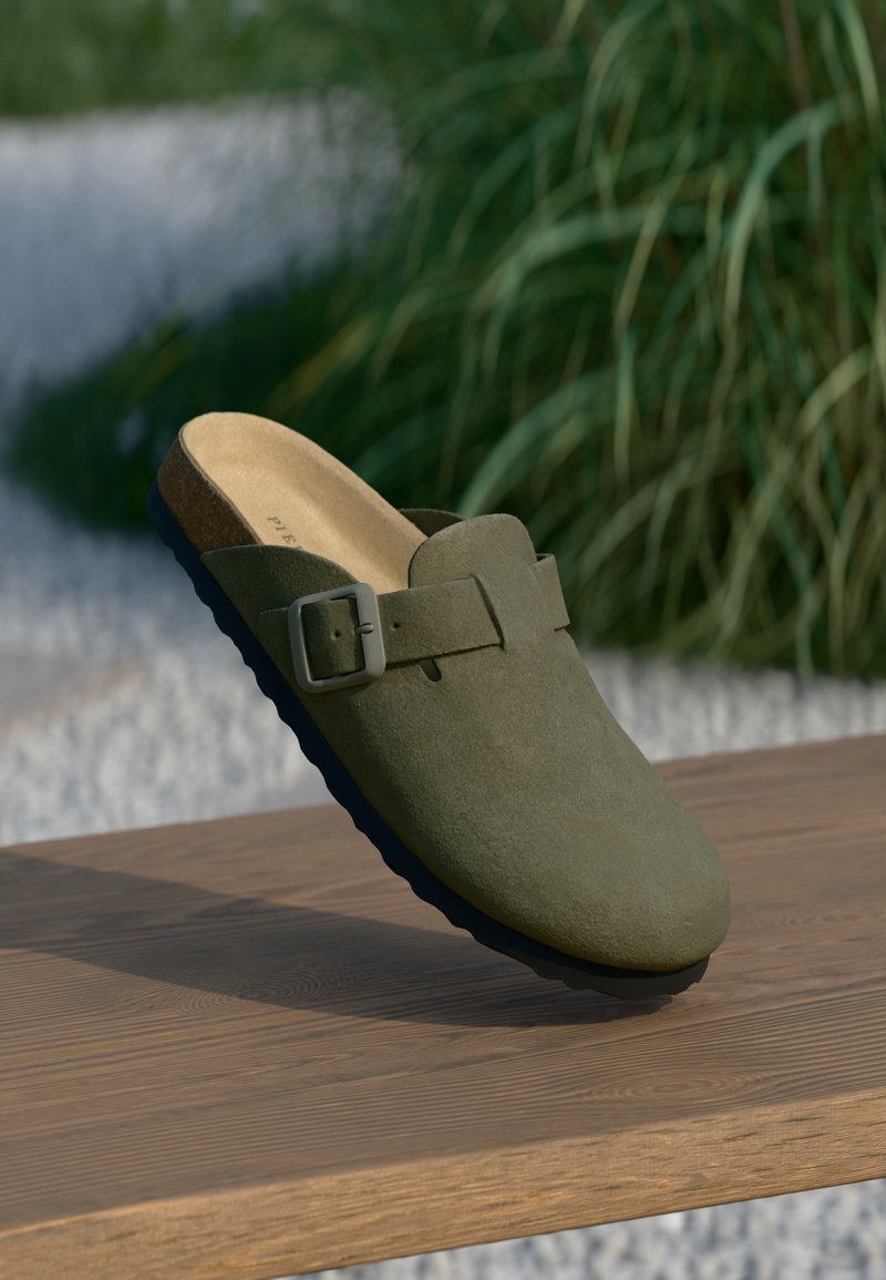 Olijfgroene suède instapklomp met verstelbare gespsluiting op houten oppervlak, buitenomgeving met vervaagde groene achtergrond en witte gravel.