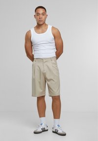 Beige geplooide shorts met een knoopsluiting, gecombineerd met een witte tanktop. Model staat met de armen achter de rug, en draagt witte en zwarte sneakers.