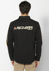 Camisa negra de manga larga con cuello puntiagudo, que presenta un gran logo de "RIDERS" en amarillo en la parte posterior y gráficos adicionales en las mangas.