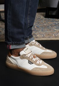 White and beige sneakers with perforated accents, textured suede, flat laces, and a rubber sole, complemented by dark denim with colorful stitching.