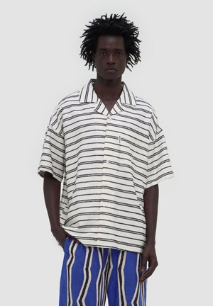 Mężczyzna o ciemnej karnacji i kręconych włosach, ubrany w białą koszulę z krótkim rękawem w czarne paski i niebieskie spodnie w paski, stoi z jedną ręką w kieszeni.