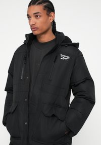 Jachetă pufă neagră cu glugă, echipată cu buzunare cu fermoar, manșoane ribbate și un logo Reebok proeminent pe piept. Textură netedă și design matlasat.