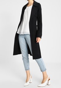 Zwarte trenchcoat met een kraag, over een witte blouse met stippen, gecombineerd met lichtblauwe broek met omslagen en witte blokhak schoenen.