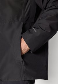Jachetă neagră impermeabilă cu un exterior texturat, buzunar lateral și detaliu "DRY VENT" pe mânecă. Fit ajustat cu un design modern.
