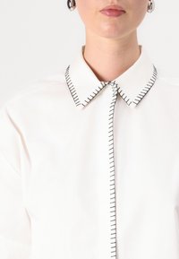 Camicia bianca con colletto caratterizzata da cuciture di contrasto nere lungo il colletto e la pattina dei bottoni. Tessuto liscio e leggero con una vestibilità strutturata.