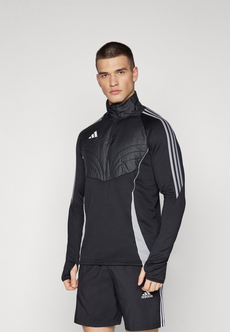Haut de sport noir à manches longues avec un panneau texturé sur la poitrine, des accents gris sur les côtés et trois bandes blanches sur chaque épaule. Porté avec un short noir.