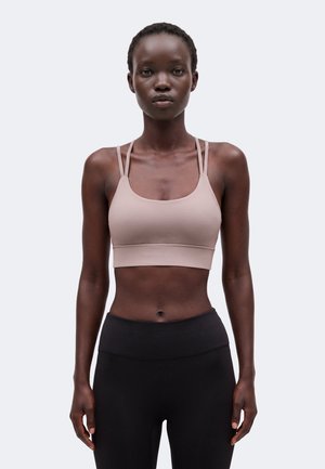 Jonge vrouw met een donkere huid die een beige sportbeha met dubbele dunne bandjes en zwarte hoge taille legging draagt, staand tegen een effen achtergrond.