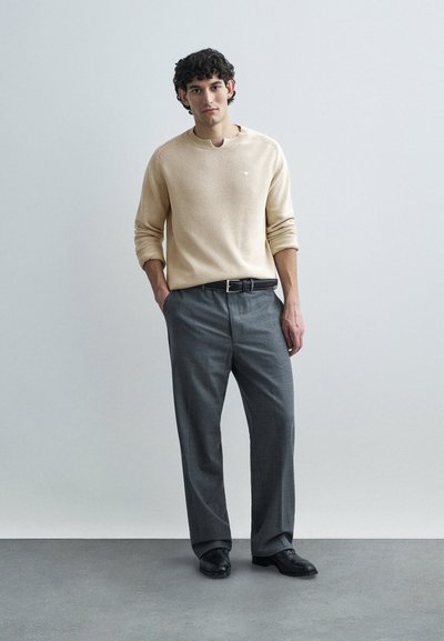 Un jeune homme se tient les mains dans les poches, portant un pull beige, un pantalon habillé gris, une ceinture noire et des chaussures noires, devant un fond uni gris clair.