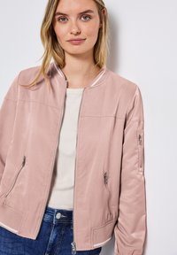 Veste bombardier rose clair avec fermeture éclair à l'avant, poches latérales et taille côtelée. Dispose d'un col simple et d'une texture lisse et brillante.