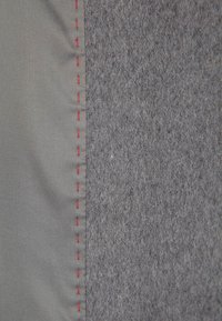 Harmaa tekstiilimateriaali, jolla on vasemmalla sileä, kiiltävä pinta ja oikealla pehmeä, teksturoitu pinta, reunassa punaiset ompeleet.