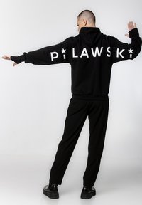 Czarna bluza z kapturem z białym napisem "PILAWSKI" na plecach. Miękka tkanina, luźny krój. Zestawiona z czarnymi spodniami i masywnymi czarnymi butami.
