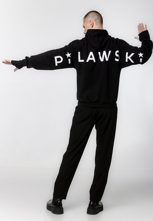 Czarna bluza z kapturem z białym napisem "PILAWSKI" na plecach. Miękka tkanina, luźny krój. Zestawiona z czarnymi spodniami i masywnymi czarnymi butami.