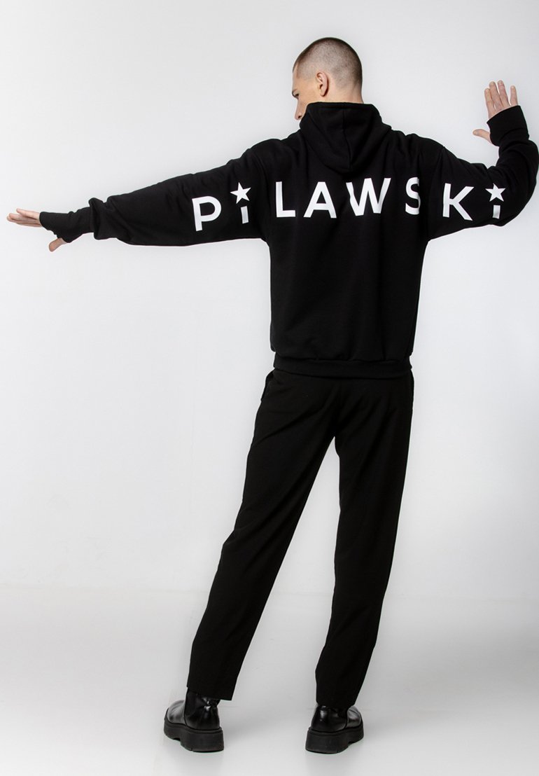 Czarna bluza z kapturem z białym napisem "PILAWSKI" na plecach. Miękka tkanina, luźny krój. Zestawiona z czarnymi spodniami i masywnymi czarnymi butami.