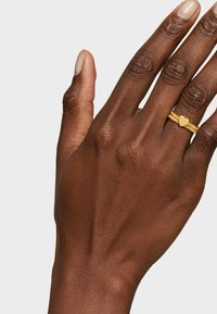Gold-toned ring with a heart-shaped accent, featuring a textured band. The hand displays a light brown skin tone.