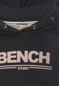 Sweat à capuche noir avec texte rose "BENCH" au-dessus d'une double ligne et mot plus petit "STUDIO" en dessous des lignes à l'avant.