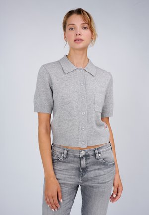 Frau trägt einen kurzärmeligen grauen Strickpullover mit Knöpfen, Kragen und einer Brusttasche, kombiniert mit hellgrauen, ausgewaschenen Jeans vor einem schlichten Hintergrund.