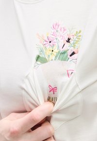 Χέρι που τραβάει το ύφασμα ενός λευκού πουκαμίσου, αποκαλύπτοντας κεντημένο πολύχρωμο μπουκέτο λουλουδιών δεμένο με ροζ φιόγκο.