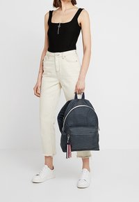 Marinblå rutig tygryggsäck med rundad form, framfack med dragkedja och dekorativ frans i rand. Modellen bär en svart topp och krämfärgade byxor.