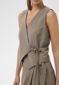 Brezrokavnik v stilu zapenjanja z nagubanim, taupe tkanino. Ima pas z metalnim okovjem in kotno linijo izreza. Gladek tekstura s strukturiranim dizajnom.
