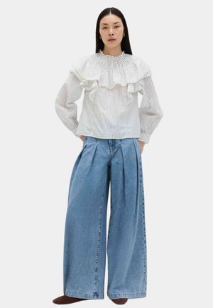 Mujer con una blusa blanca con volantes y finas rayas, y pantalones vaqueros azules plisados de pierna ancha, de pie con las manos en los bolsillos sobre un fondo liso.