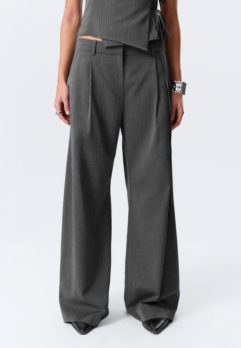 Pantalon large à rayures grises avec plis, doté d'une taille ajustée. Tissu lisse avec une forme structurée. Chaussures noires à bouts pointus visibles.