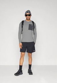 Γκρι μακρυμάνικο πουκάμισο με τσέπη, μαύρο σορτς, μαύρα αθλητικά παπούτσια και γυαλιά ηλίου. Το μοντέλο φορά γκρι σκουφάκι και μαύρο σακίδιο πλάτης.