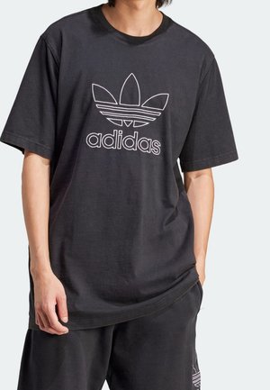 Osoba nosząca czarną koszulkę Adidas z białym logo i pasujące czarne spodnie, stoi z jedną ręką w kieszeni.