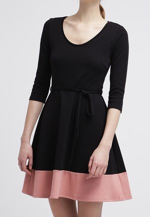 Femme portant une robe noire à manches trois-quarts, une ceinture nouée à la taille et un large ourlet rose sur la jupe près du milieu de la cuisse.