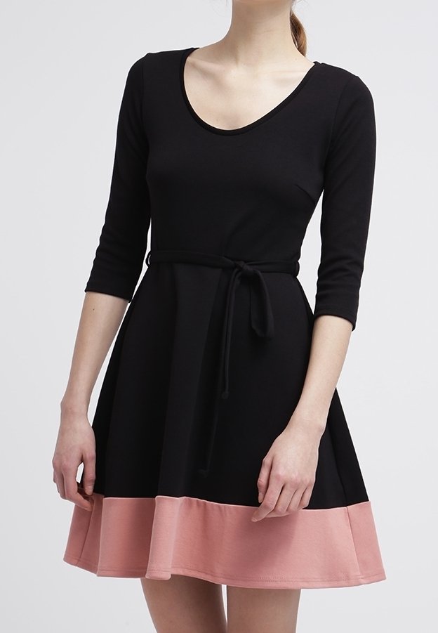 Femme portant une robe noire à manches trois-quarts, une ceinture nouée à la taille et un large ourlet rose sur la jupe près du milieu de la cuisse.