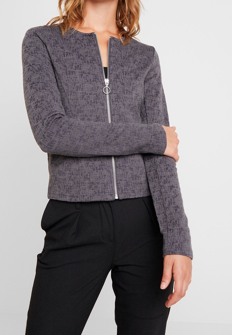 Femme portant une veste zippée gris à motif avec tirette anneau et pantalon noir, debout les bras croisés devant un fond blanc.