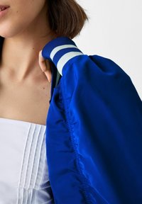 Modrá bomber bunda s lesklým povrchem, se žebrovanými manžetami v bílé a námořnické modři. Bunda má na límci záhybové detaily.